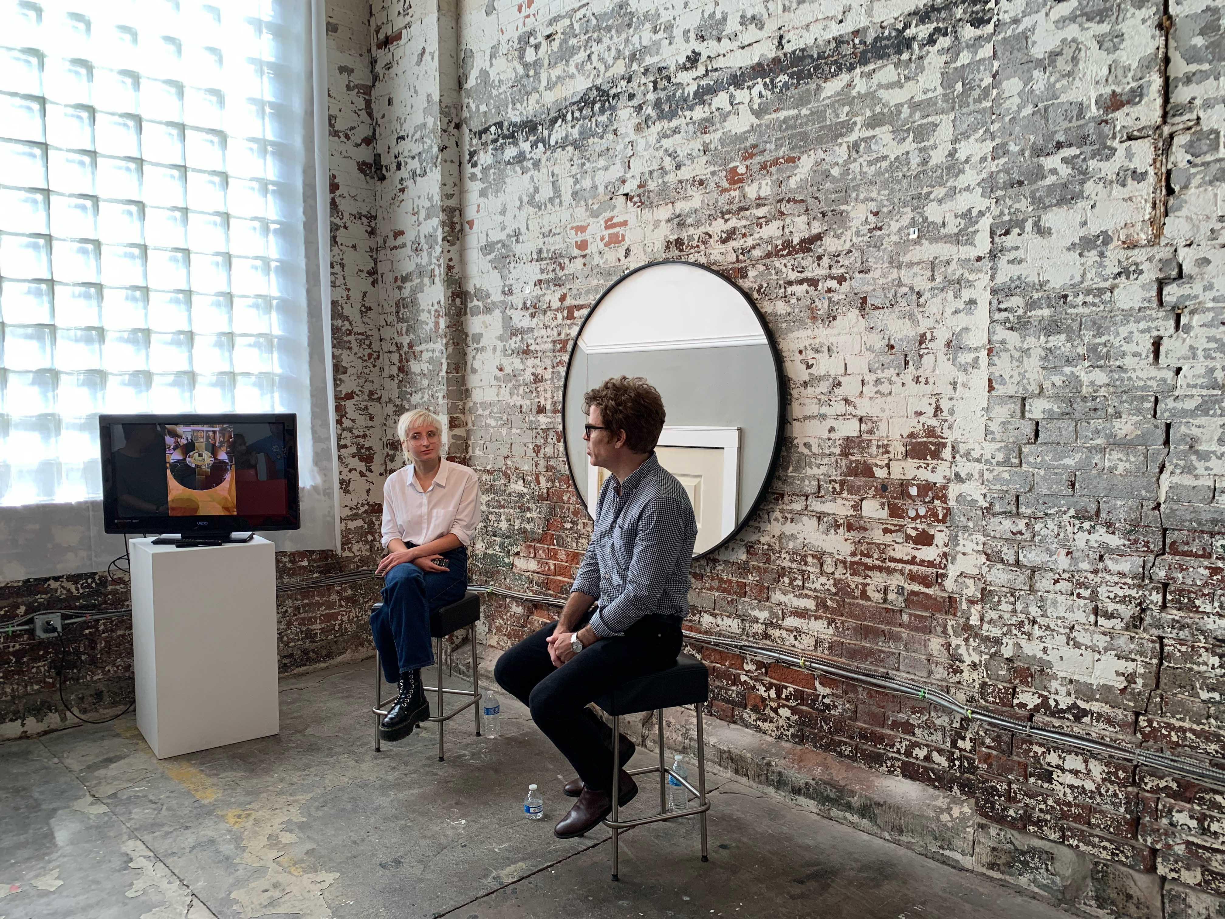two people sit in front of a red brick wall with a monitor on a pedestal beside them 